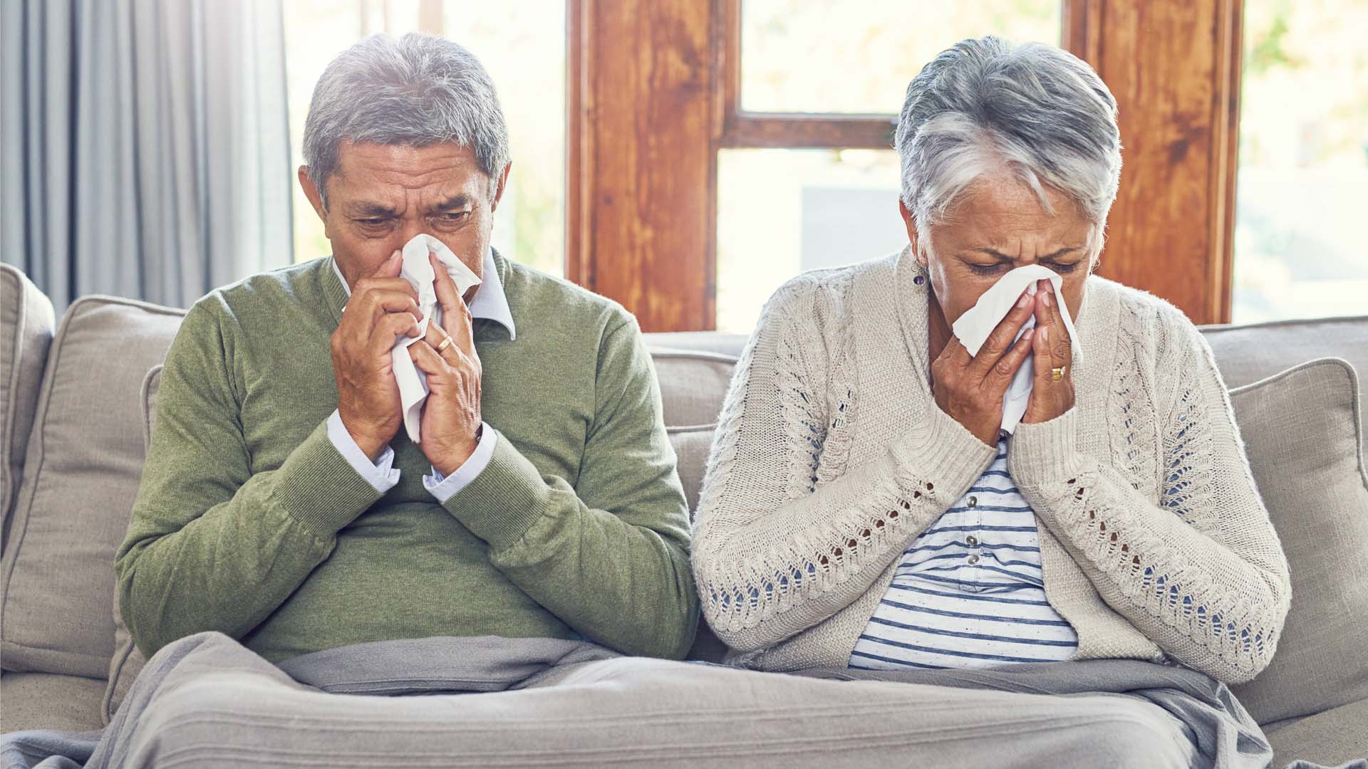 A senior couple with colds blowing their noses while siting on a couch under a blanket