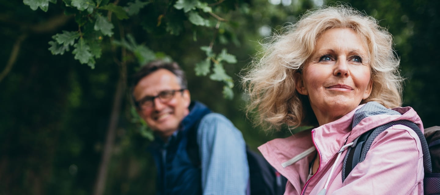 A blonde woman wears a pink coat and backpack on a hike outdoors with a male partner