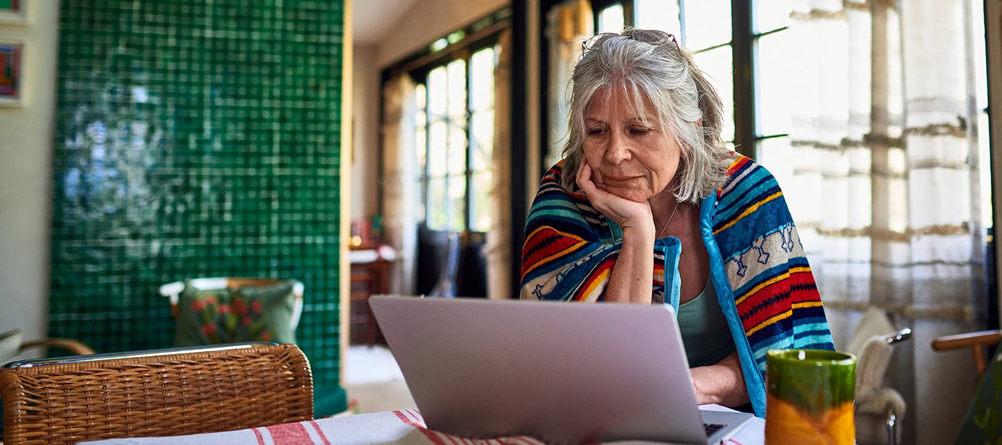 A woman wrapped in blanket standing at dining table looking at her laptop.