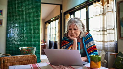 A woman wrapped in blanket standing at dining table looking at her laptop.