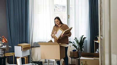 A woman in her 30's standing in the middle of a messy room, looking in a box in front of her and holding picture frames in her hands.