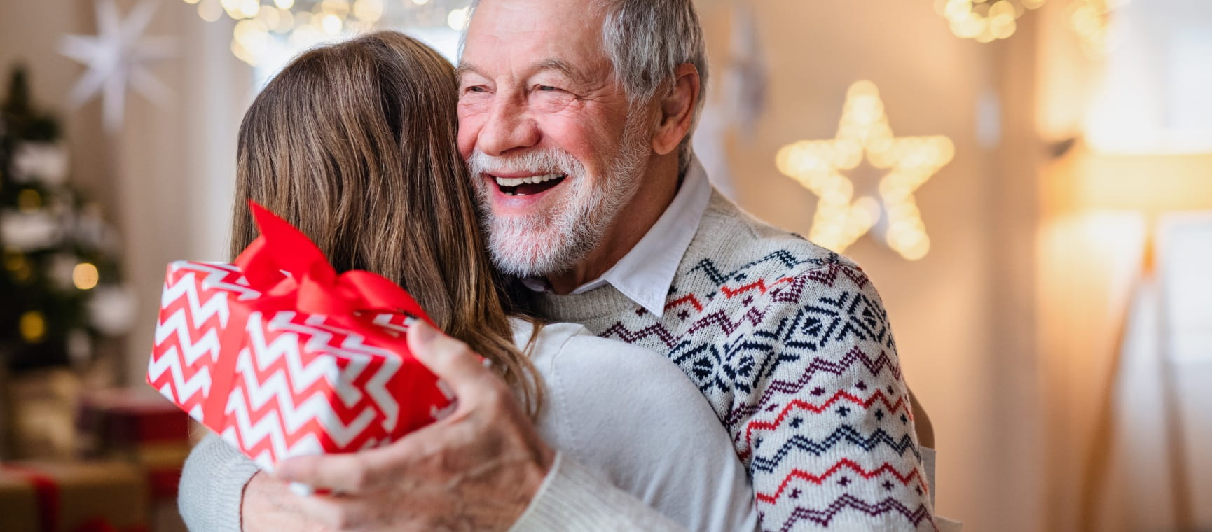 A senior man hugs a family member while holding a Christmas gifts with Christmas lights in the background