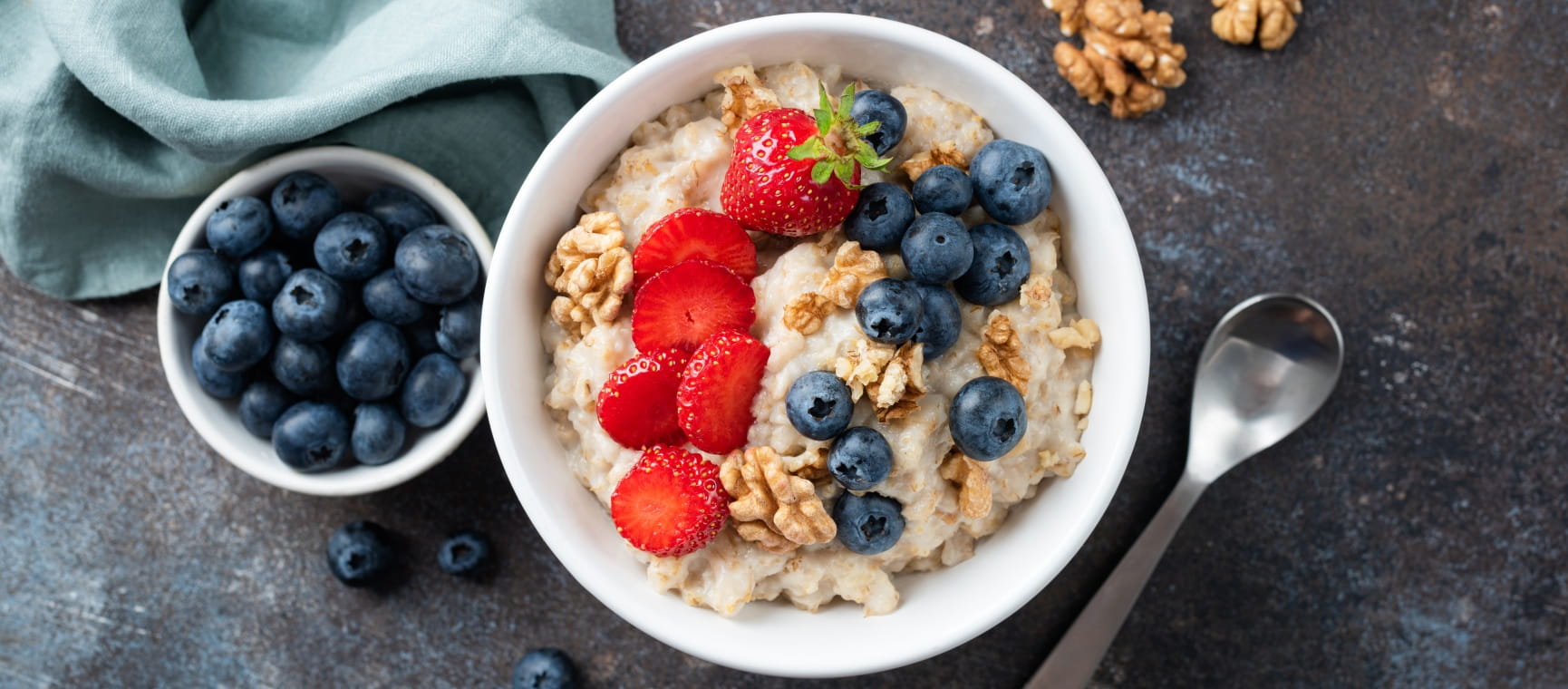 A bowl of porridge topped with blueberries, strawberries and walnuts, with a pot of blueberries on the side