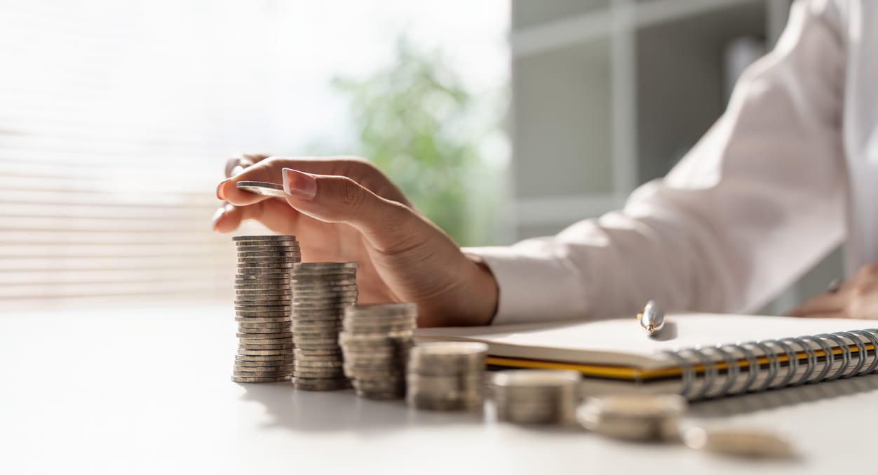 Someone with increasingly higher stacks of coins in front of them on a table with a notepad and pen