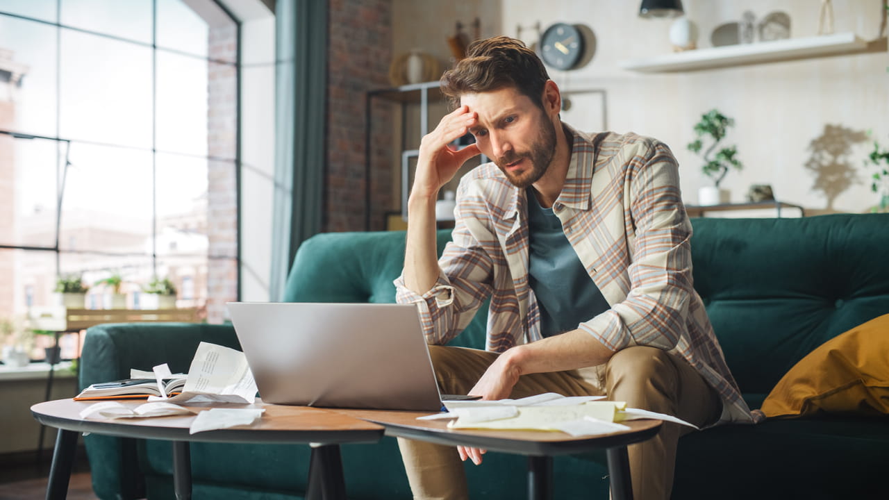 A man sat at a laptop, surrounded by paperwork, with his head in his hand in frustration