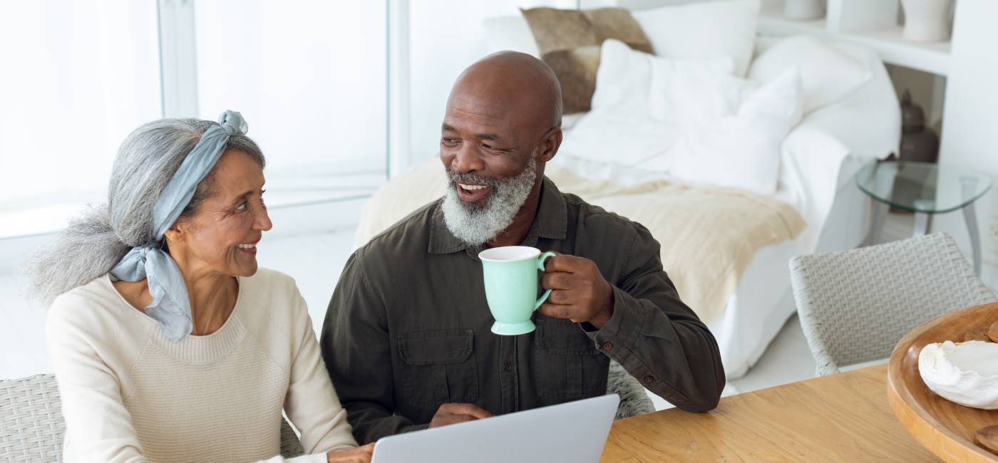 Front view of diverse senior couple using laptop on table while man holds a cup in beach house