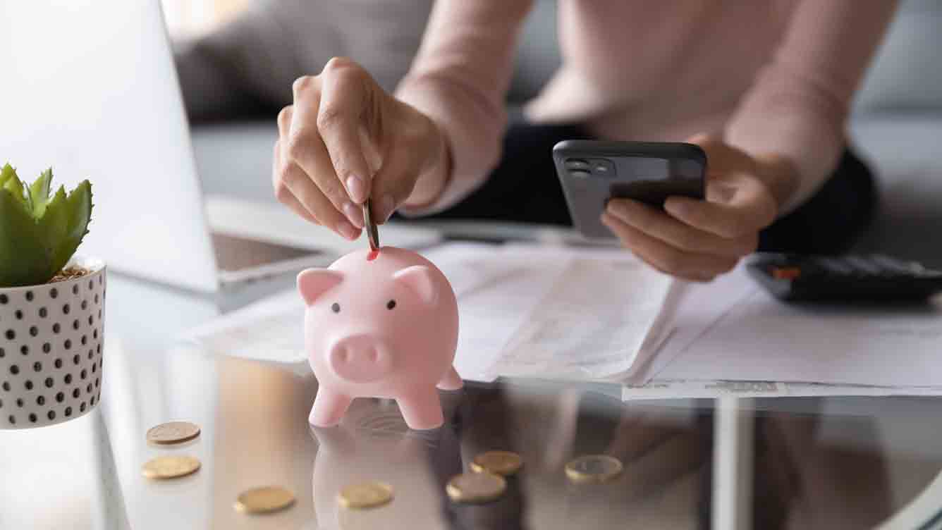 A woman sat at a desk with a calculator and putting money into a piggy bank